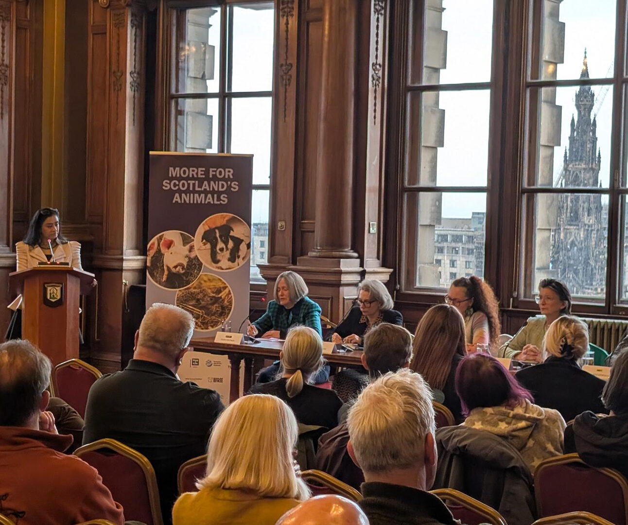 A room full of people seated, facing a panel of five people at the front of the room