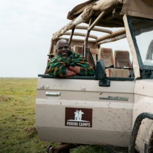 Photograph of a person wrapped in a colorful Maasai shawl sitting inside a beige open-top safari vehicle labeled "Porini Camps" on a grassy plain. The vehicle's interior shows multiple empty seats, indicating a safari or wildlife tour setting in a natural environment.