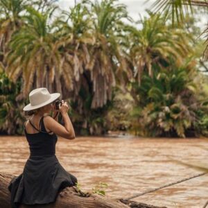 Photograph of a woman wearing a white hat and black dress sitting on a log by a muddy river, taking a photo with a camera. Dense green palm trees form a lush background, highlighting a natural, tropical riverside setting.