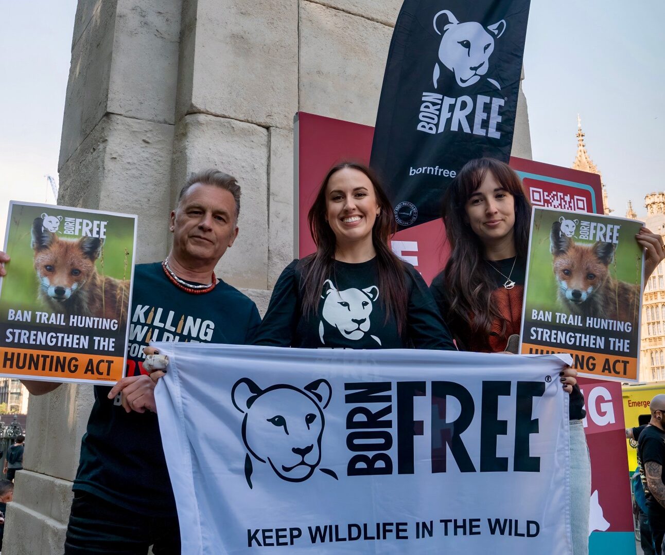 Photograph of a group of protesters including Chris Packham, Olivia Evand and Megan McCubbin outside a historic building holding signs and banners advocating to ban trail hunting and strengthen the Hunting Act. Signs feature a white bear logo with 