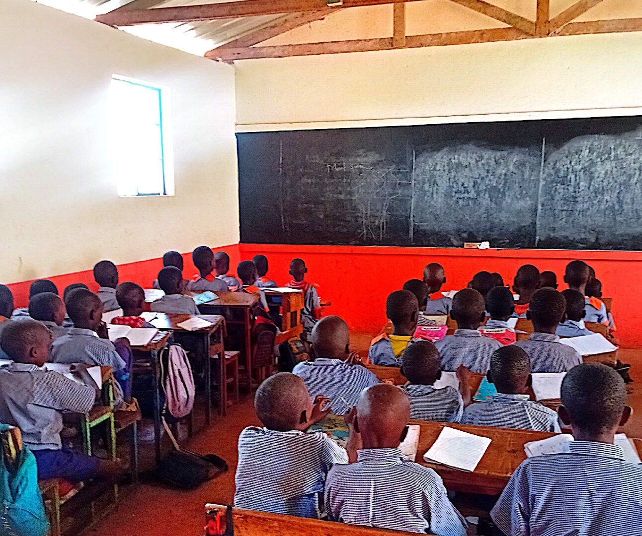 Photograph of a classroom in Kenya filled with students seated at wooden desks facing a large blackboard. The room has high windows allowing natural light, and students appear attentive, wearing matching uniforms.