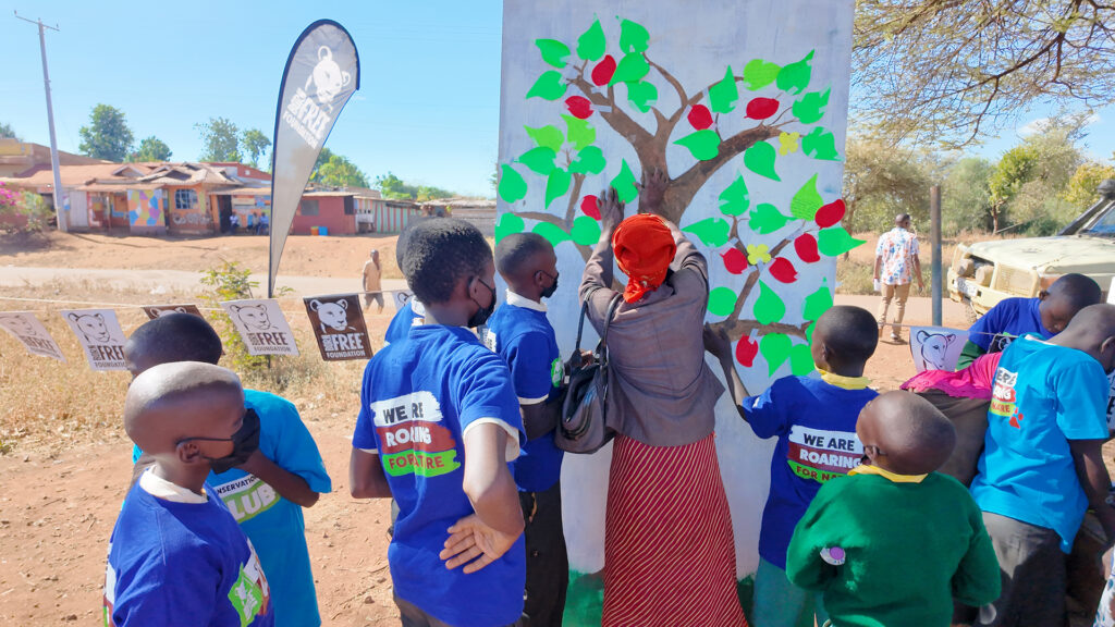 A group of children in matching blue Born Free tshirts are stood around a large painting of a tree, with leaves containing written messages stuck to it. A woman is at the centre, placing a leaf on the tree.