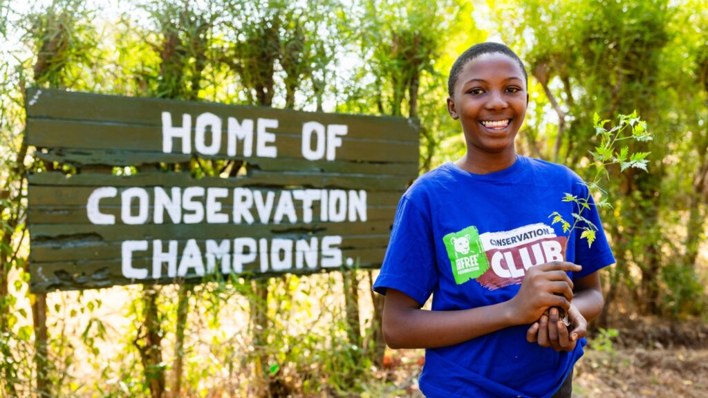 Photograph of a smiling young person holding a small plant in front of a wooden sign that reads "HOME OF CONSERVATION CHAMPIONS." The setting is outdoors with greenery, highlighting a conservation or environmental club promoting tree planting and ecological awareness.