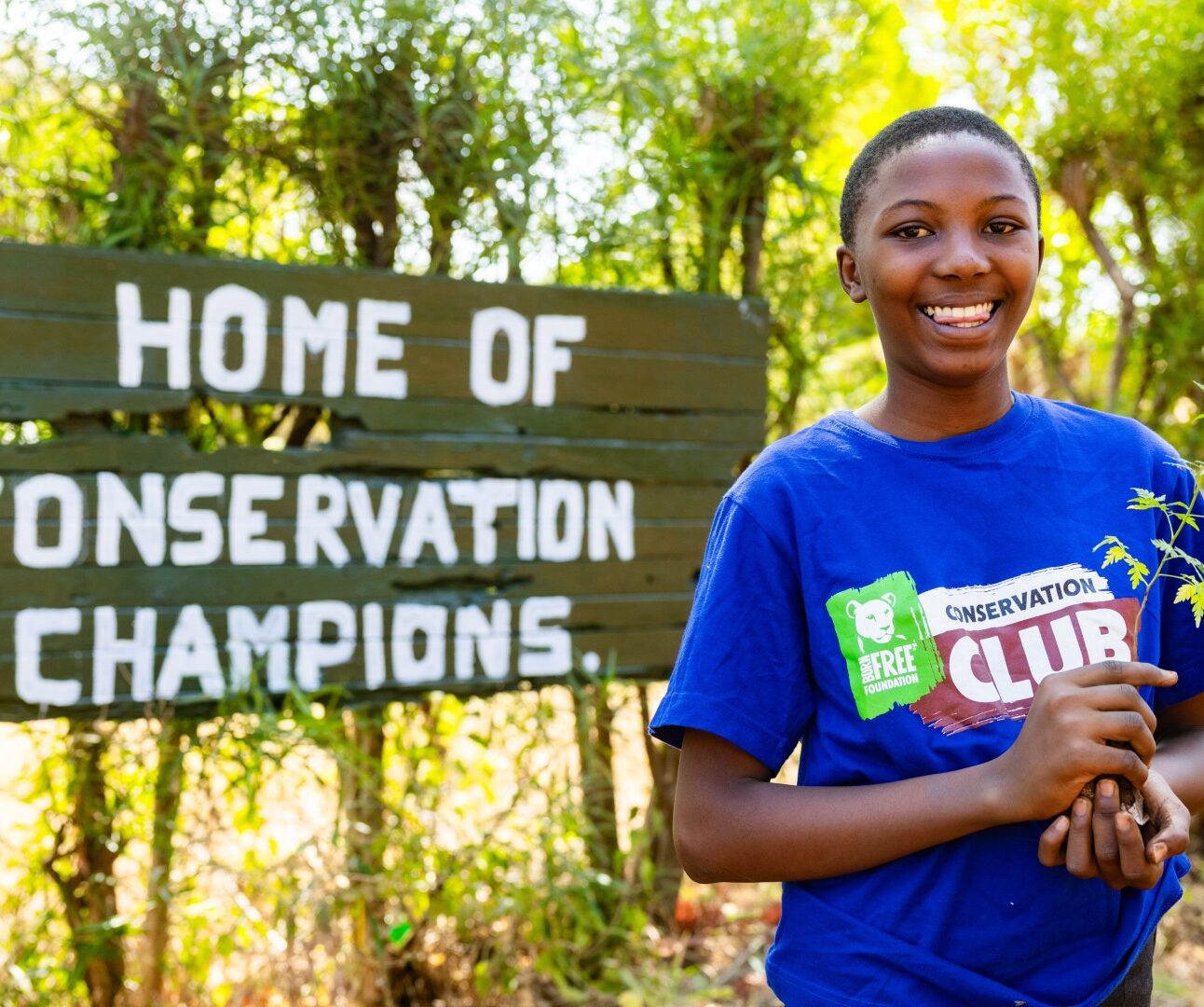 Photograph of a smiling young person holding a small plant in front of a wooden sign that reads 