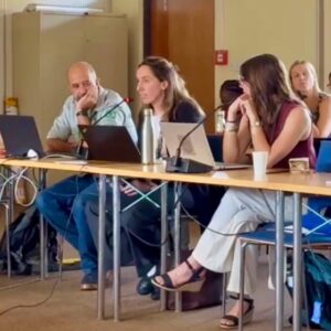 Photograph of a group meeting with four people seated at a long table using laptops and microphones, suggesting a discussion or presentation setting. The room features neutral tones, with participants focused on conversation, highlighting collaboration and engagement.