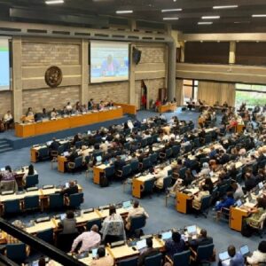 Photograph of a large conference hall filled with numerous attendees seated at desks with laptops, facing a panel of speakers at the front. The room features a large emblem on a brick wall and a projection screen displaying a speaker, indicating a formal meeting or legislative session.