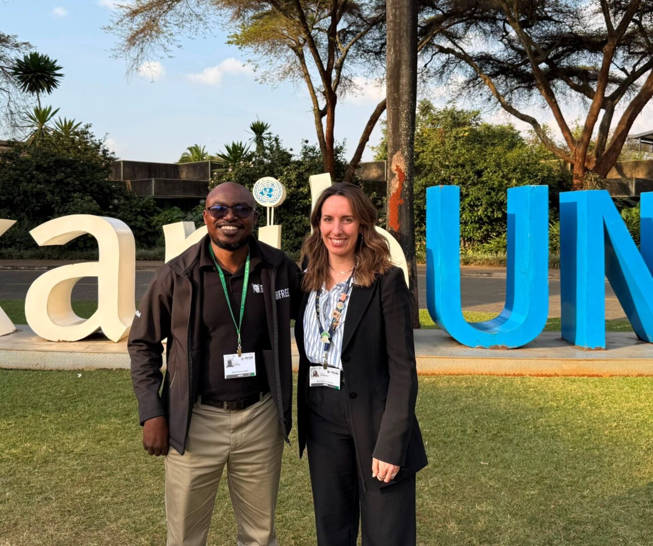 Photograph of two people standing on grass in front of large, colourful letters outdoors with trees and buildings in background. Both individuals wear conference badges, suggesting attendance at an event or university-related gathering.