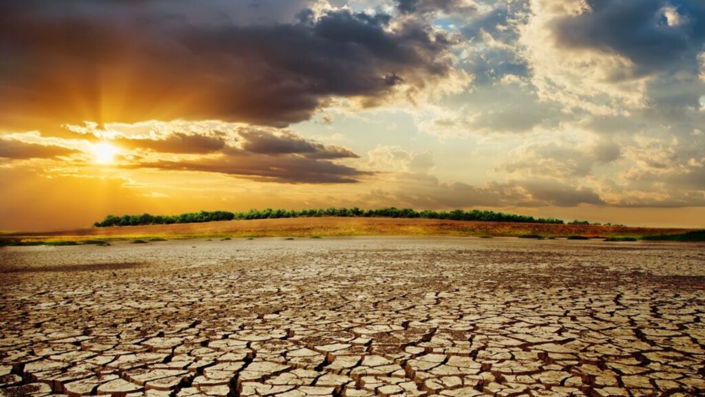 Photograph of a cracked, dry landscape under a dramatic sky with scattered clouds and sunlight breaking through. The scene highlights drought conditions with parched earth stretching toward a distant green tree line on the horizon.