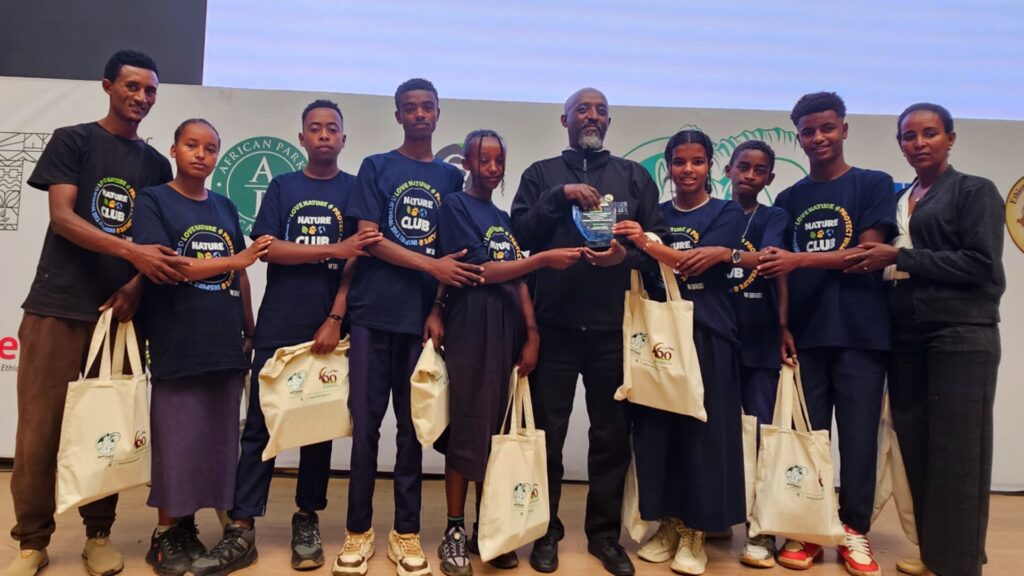 Photograph of a group of people standing in a row on stage, all wearing matching navy blue "Nature Club" t-shirts and holding beige tote bags with logos. The setting appears to be an event or award ceremony focused on environmental or nature-related activities, highlighted by the group's uniform attire and presentation.