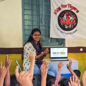 Photograph of a woman sitting on a desk in front of a laptop displaying "Kids for Tigers" logo, engaging with a group of children raising their hands. A banner behind her features a tiger face and text "Kids for Tigers" and "The Sanctuary Tiger Programme," indicating an educational session about tiger conservation.