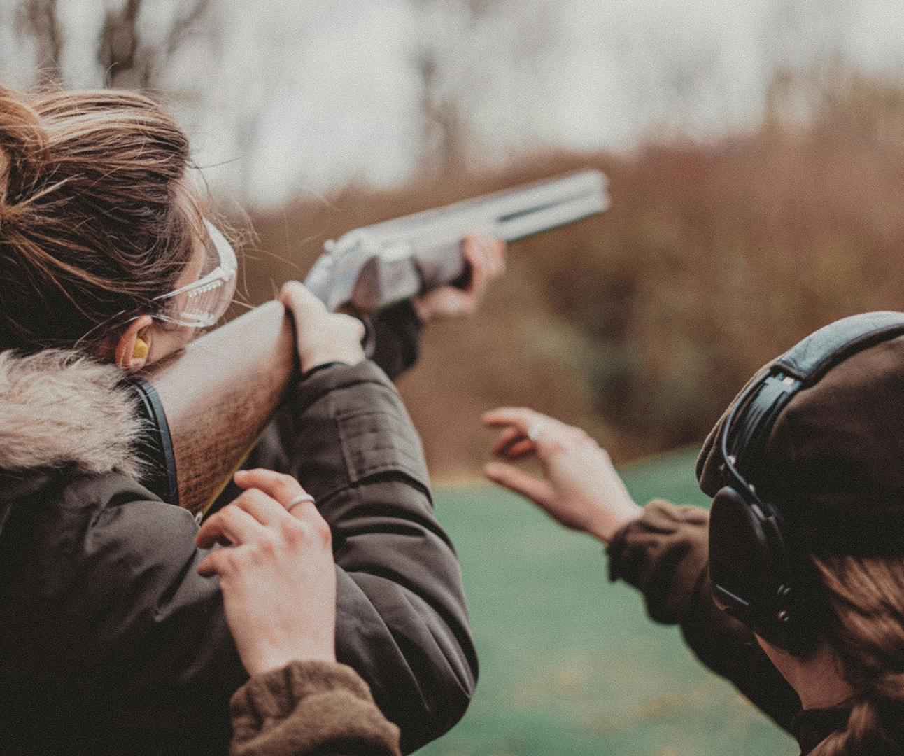 Two women are pointing guns at the sky, on what looks like a winter's day in the countryside