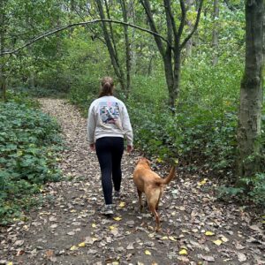 A photo of Katie Still walking through a forest with her dog. They are walking away from the camera