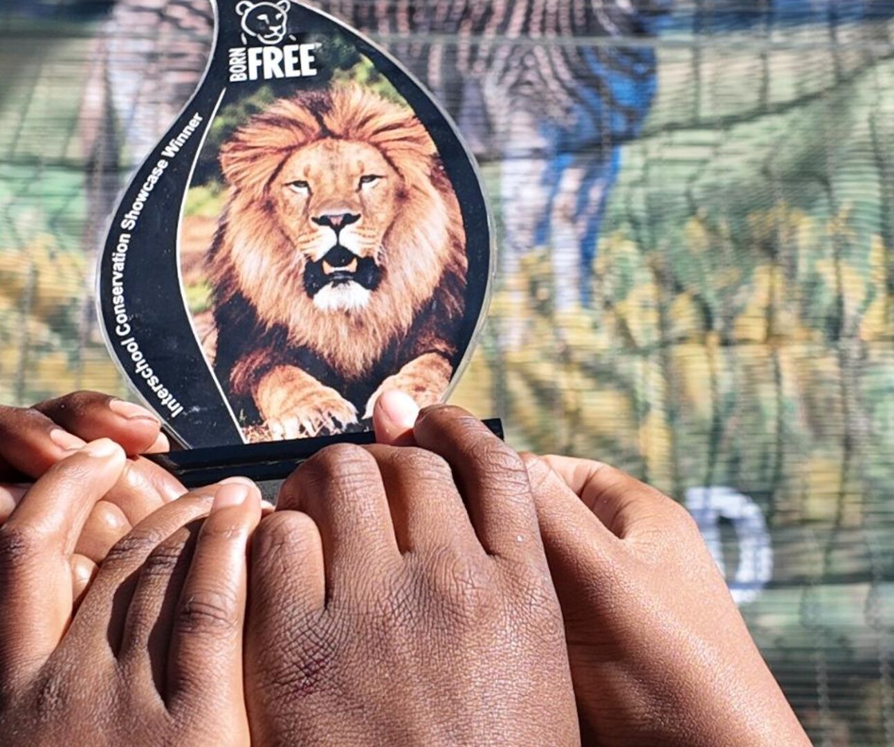 Photograph showing a group of people raising joined hands around a card featuring a lion and “BORN FREE” text, symbolizing community solidarity. A colourful mural behind them includes “Communities” lettering, animal artwork, and leafy outdoor surroundings.